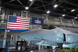 An airplane sits inside a hanger. Behind the aircraft is an American flag and a sign that reads, "Arsenal of Freedom."