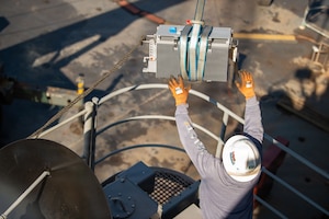 A man wearing a protective helmet and gloves reaches toward a piece of equipment suspended by a strap.