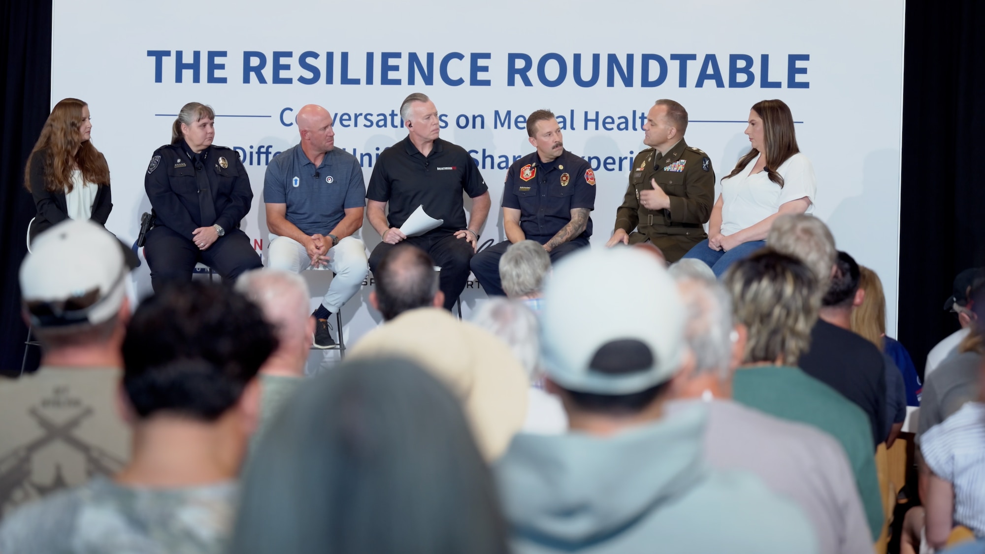 A wide shot of a panel of speakers, sitting on a stage in front of microphones inside a stadium club room.