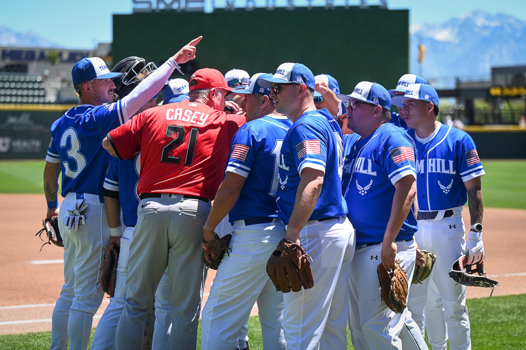 Retired Major League Baseball player Sean Casey stands in the center of a tight, circular huddle of Hill Air Force Base baseball players in blue and white uniforms, speaking to them on a green baseball field.