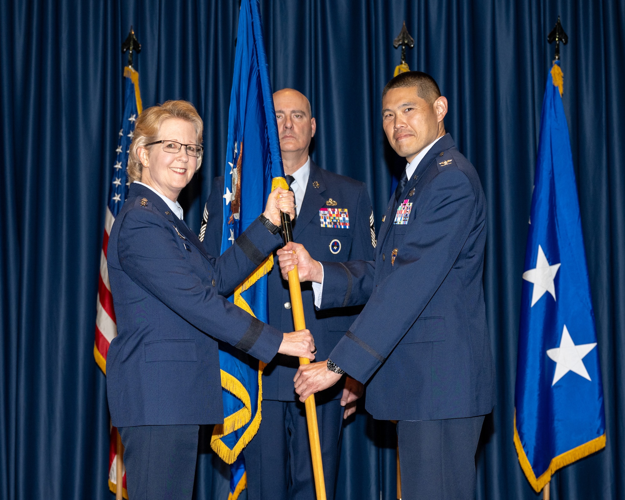 A woman and man, both wearing blue military service dress uniforms, pose for a photo while holding a flag staff and blue flag