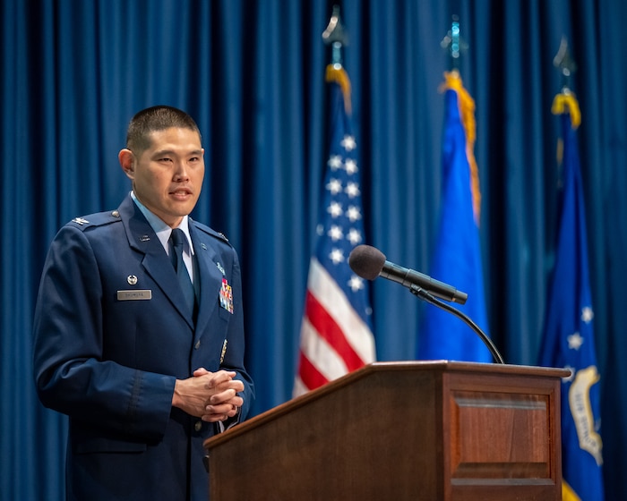A man, wearing a blue military service dress uniform, speaks behind a podium and microphone. Flags can be seen in the background in front of a blue back drop