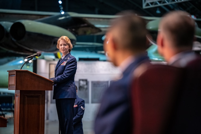 A woman, wearing a blue military service dress uniform, stand behind a podium and microphone, while looking towards two people sitting in the foreground
