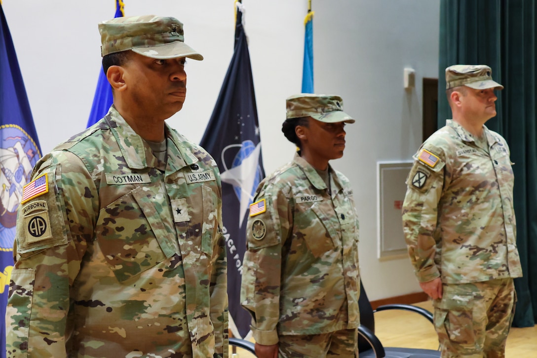 Three people wearing camouflage military uniforms stand on stage in a line.