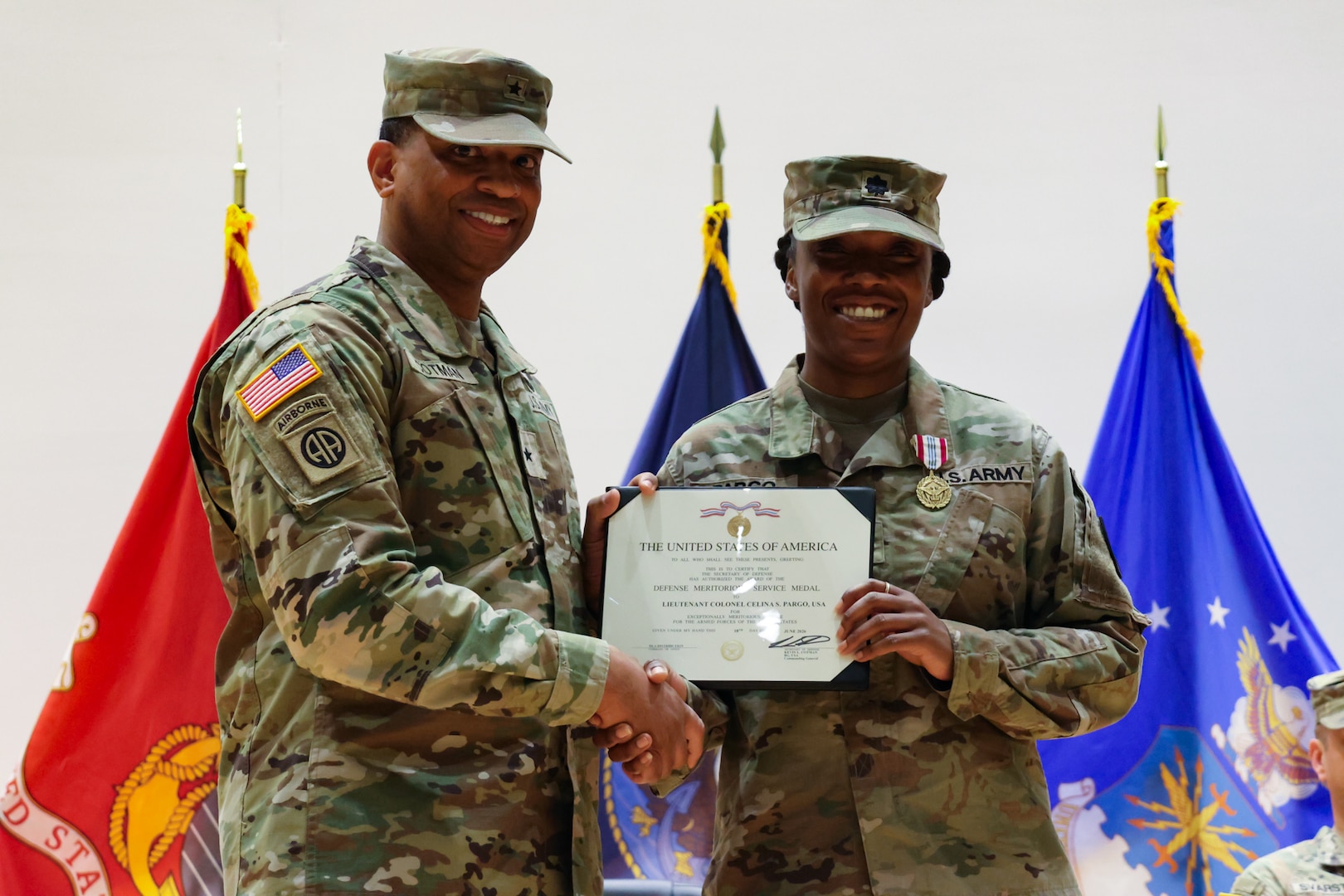 Two people wearing camouflage military uniforms shake hands and hold a certificate.
