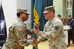 Two men wearing camouflage military uniforms pass a flag between one another