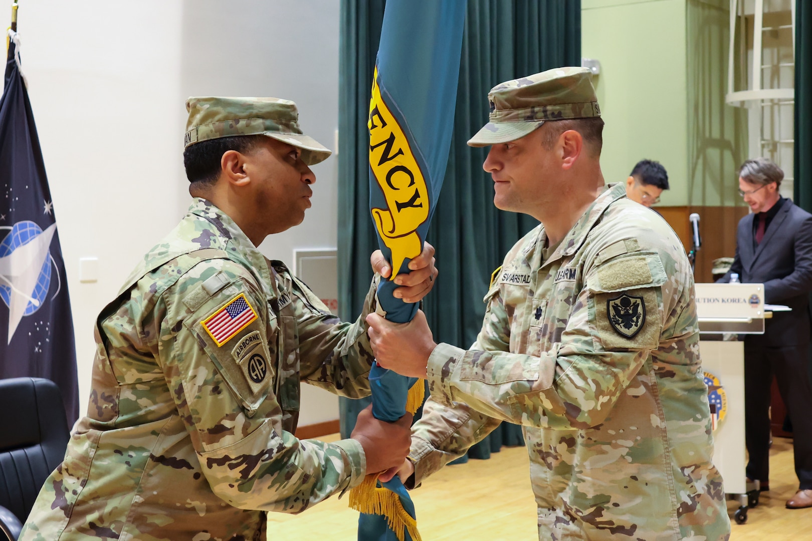 Two men wearing camouflage military uniforms pass a flag between one another