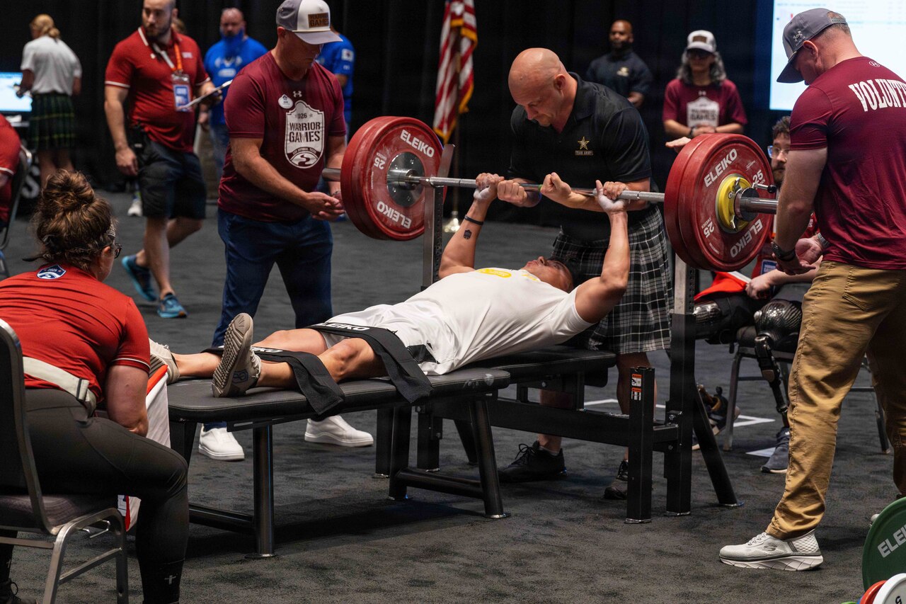 A man in athletic attire lies on a weight bench and grips a barbell, while three people in similar attire assist and a woman sits in a chair to observe; in the background, people in similar attire do similar activities.