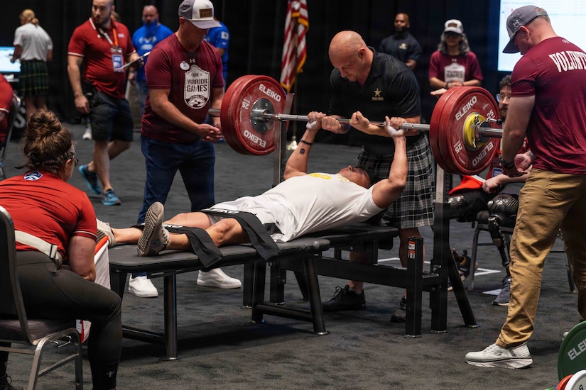 A man in athletic attire lies on a weight bench and grips a barbell, while three people in similar attire assist and a woman sits in a chair to observe; in the background, people in similar attire do similar activities.