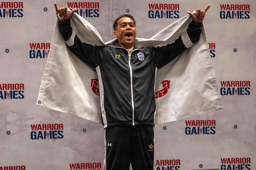 A man wearing athletic attire and an award medal lifts his arms in celebration while holding a service flag around his shoulders,