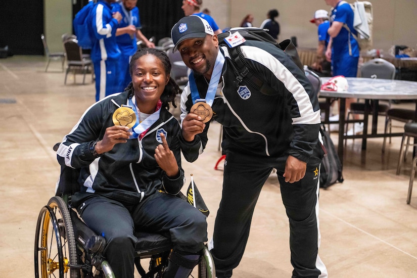 A woman in athletic attire sits in a wheelchair holding a medal around her neck while a man in similar attire leans next to her, also holding a medal, as they pose for a photo.