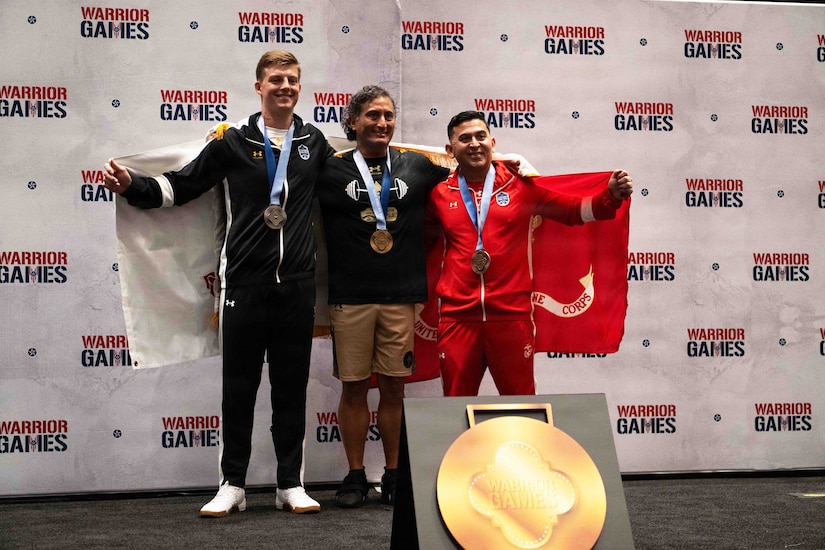 Three men wearing athletic attire and award medals hold service flags around their shoulders while standing and smiling for a photo.