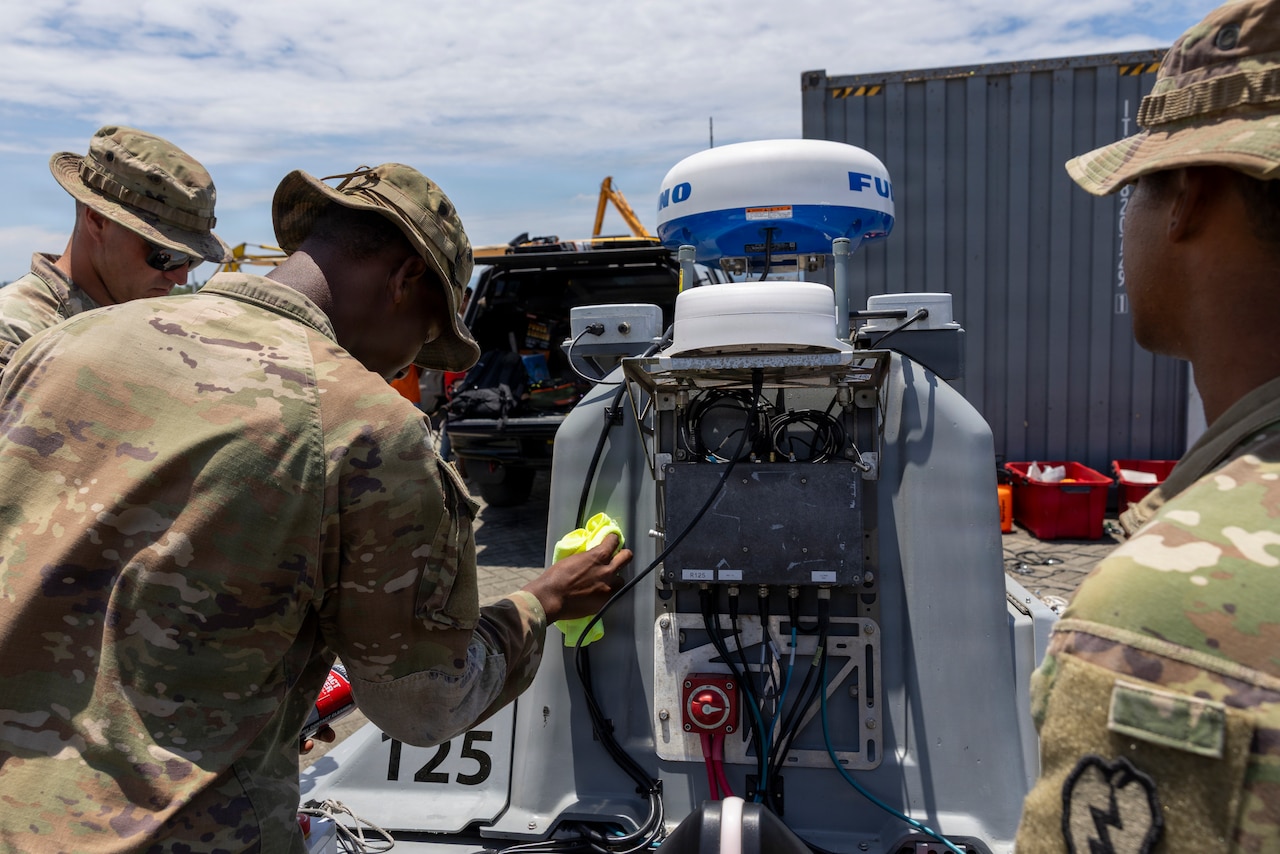 A man wearing a camouflage military uniform checks the engine on an unmanned surface vessel that is on a trailer as two other men in similar attire observe.