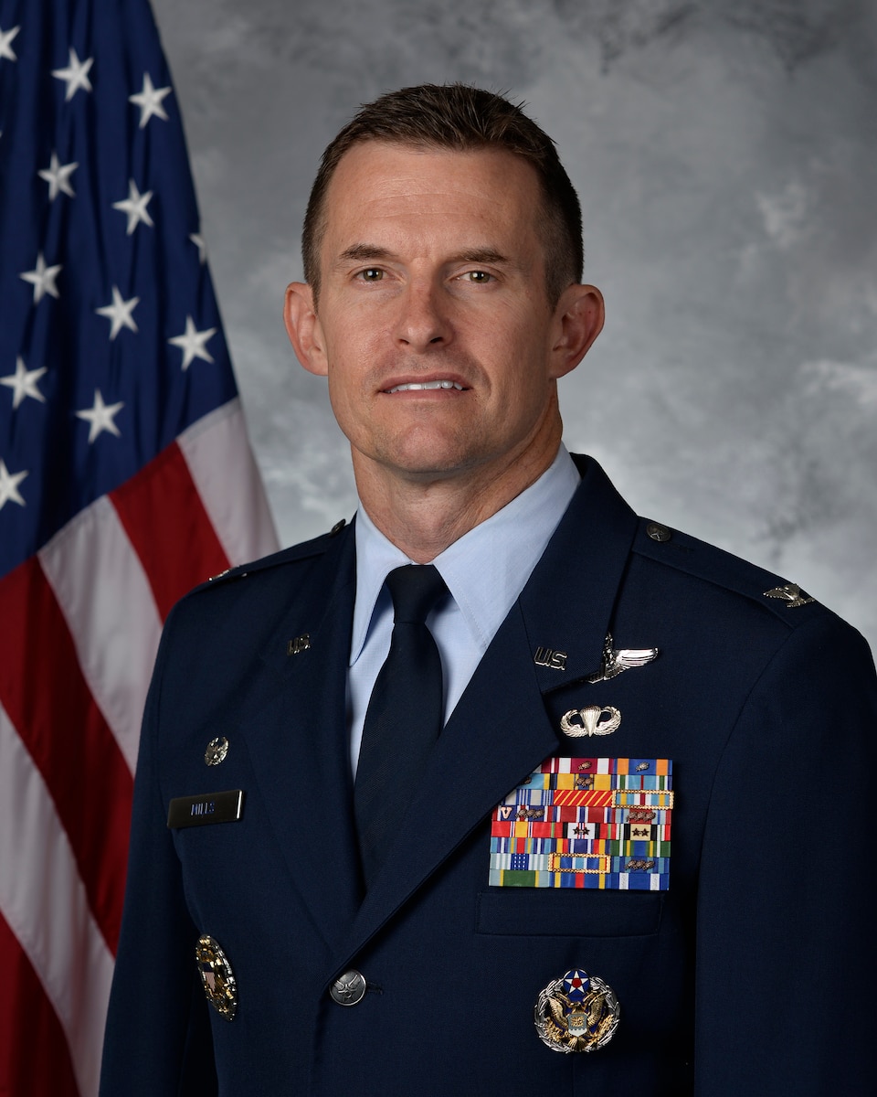 Col Mills poses for an official photo with a U.S. flag behind him with a grey background.
