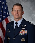 Col Mills poses for an official photo with a U.S. flag behind him with a grey background.