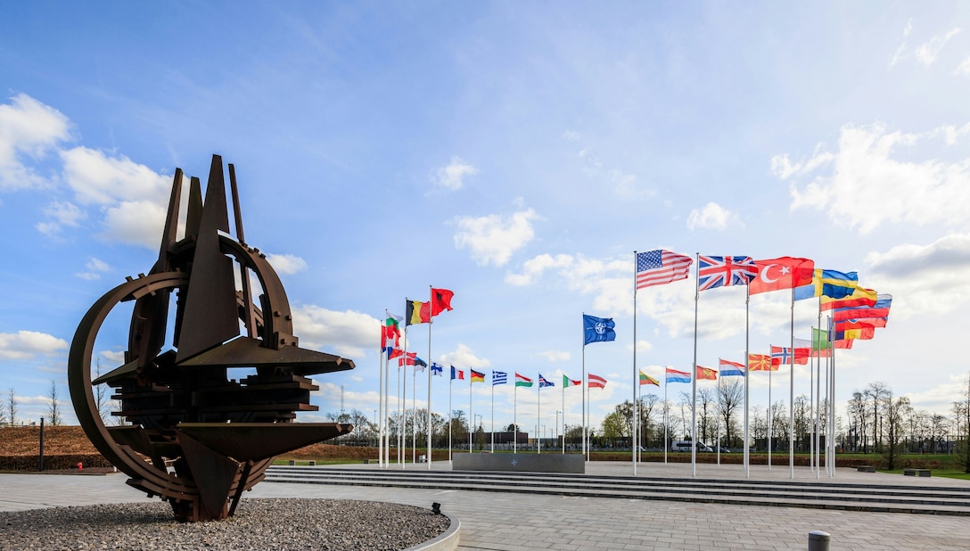 A sculpture of a stylized NATO emblem stands near a circle of national flags on a paved area.