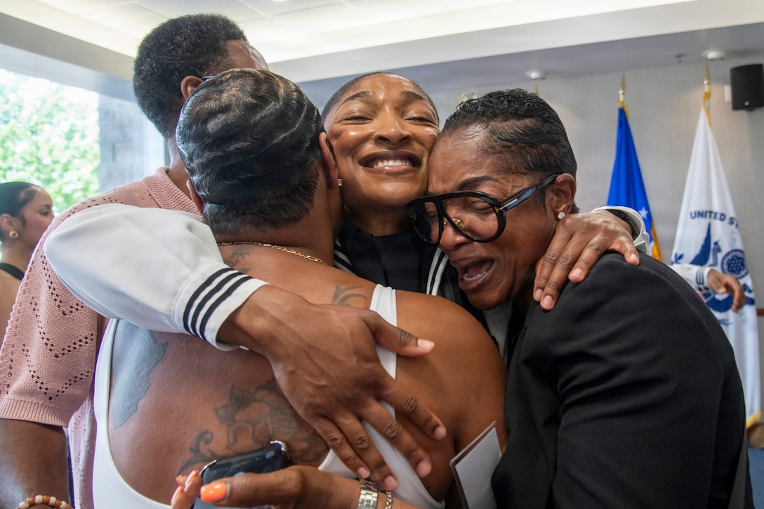 A sailor and three members of her family hug. Service flags and other people are in the background.