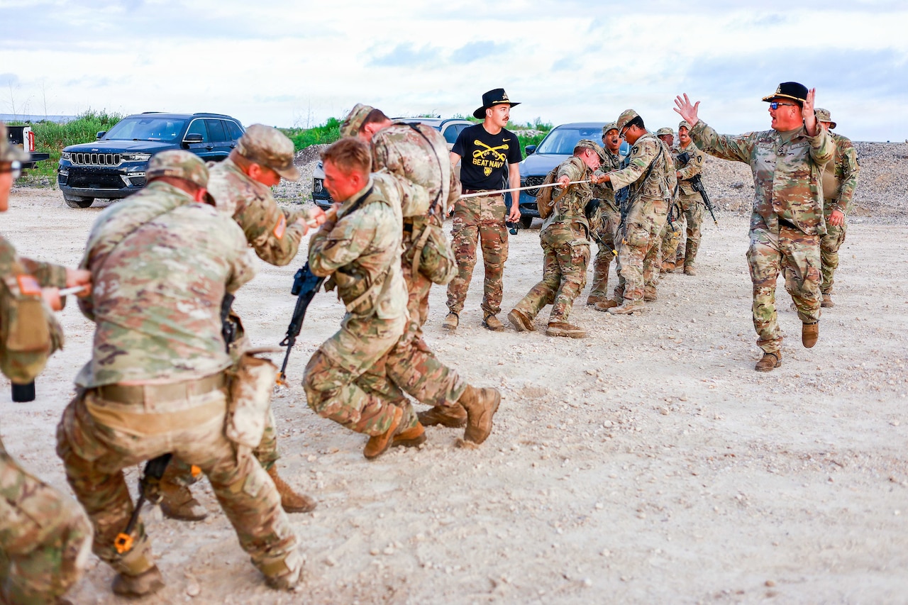 A group of soldiers conduct tug-of-war while wearing rifles, while two soldiers in cavalry hats cheer them on.