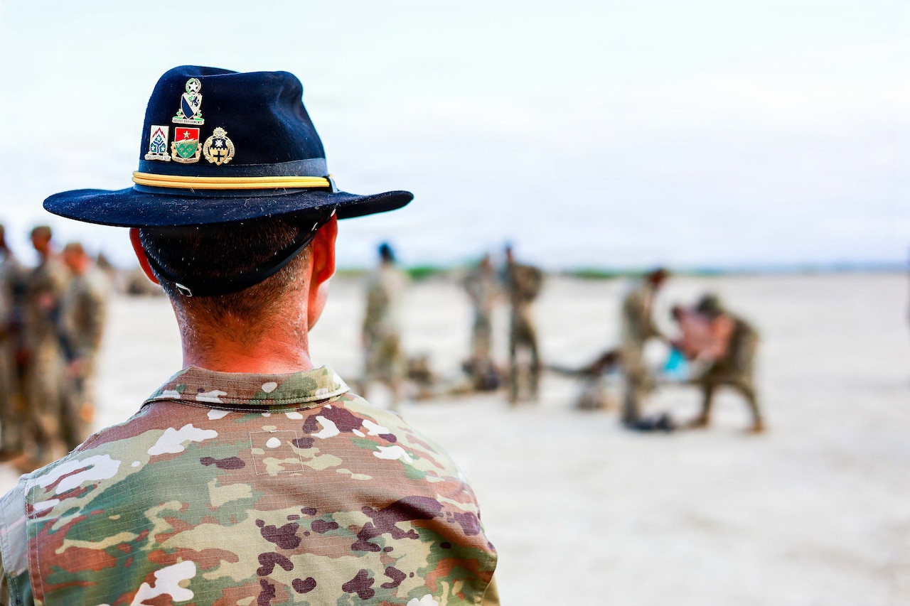 A soldier wearing a cavalry hat is  seen in the foreground with several soldiers out of focus in the distance.