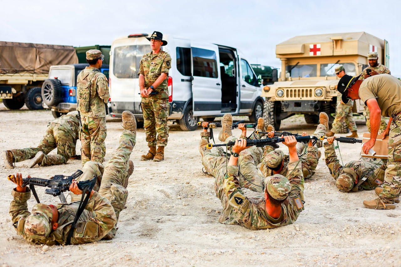 A group of soldiers balance rifles with arms and legs up while on their backs on the ground and other soldiers monitor while standing, with vehicles in the background.
