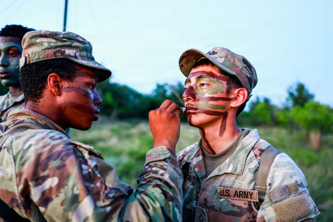 A service member applies camo paint to another's face outdoors.