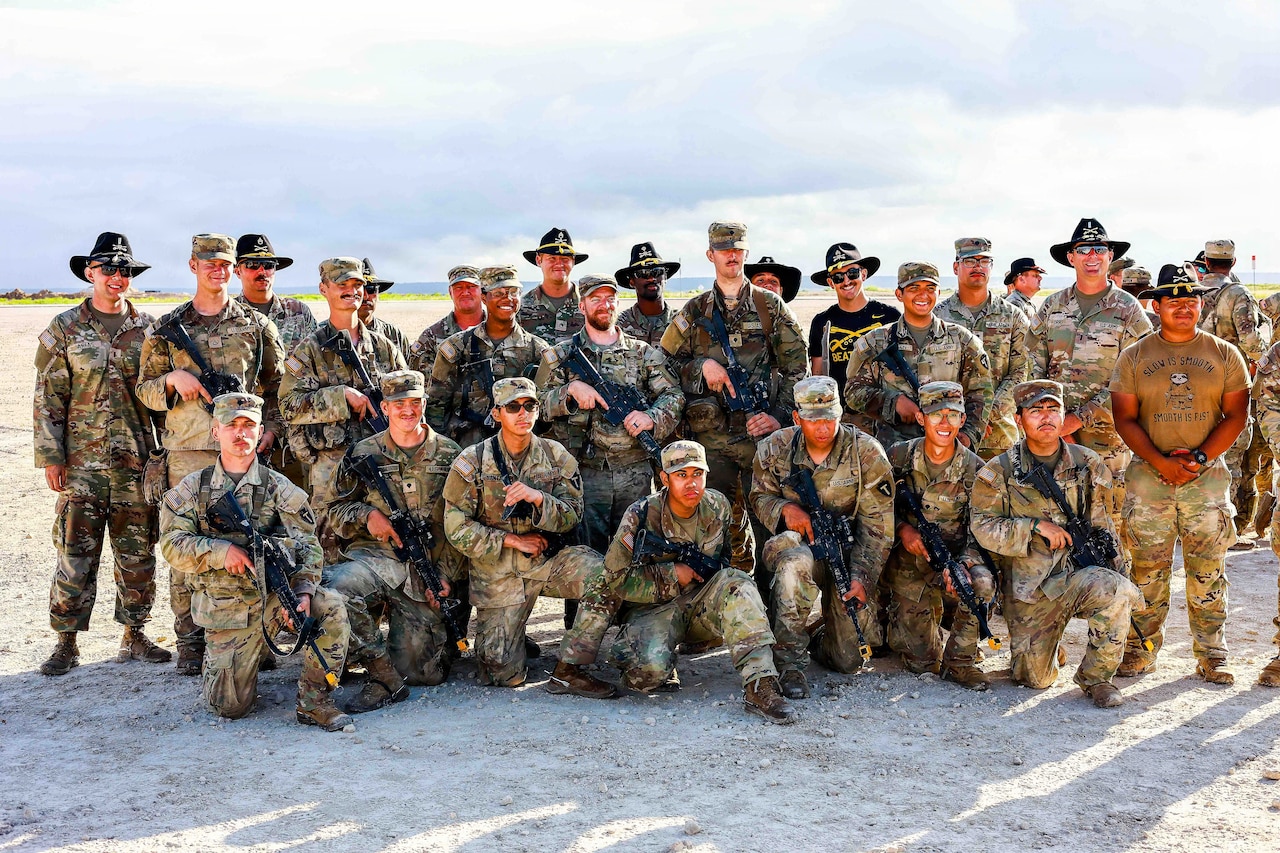 A group of soldiers, some in dark hats, stand and crouch to form two rows while looking at the camera outdoors.