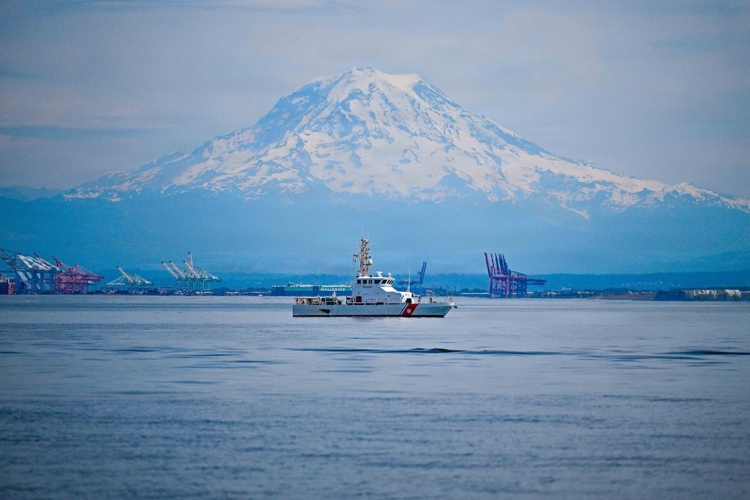 A Coast Guard cutter is in the water, with a snow-covered mountain and port equipment in the background.