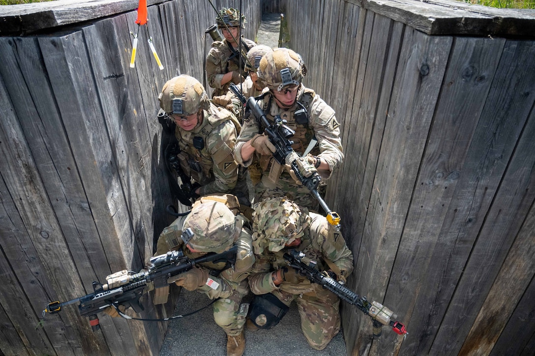 A group of six soldiers stands in a narrow space between two wooden structures, with the soldiers at the front pointing rifles and peering around the structures.