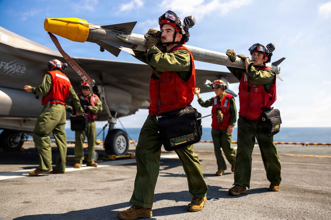Two Marines wearing green overalls, orange vests and helmets with goggles and ear muffs, carry a rocket on their shoulders, while three other Marines wearing similar clothing and gear, stand near a fighter jet aircraft on the deck of a ship with the ocean in the background.