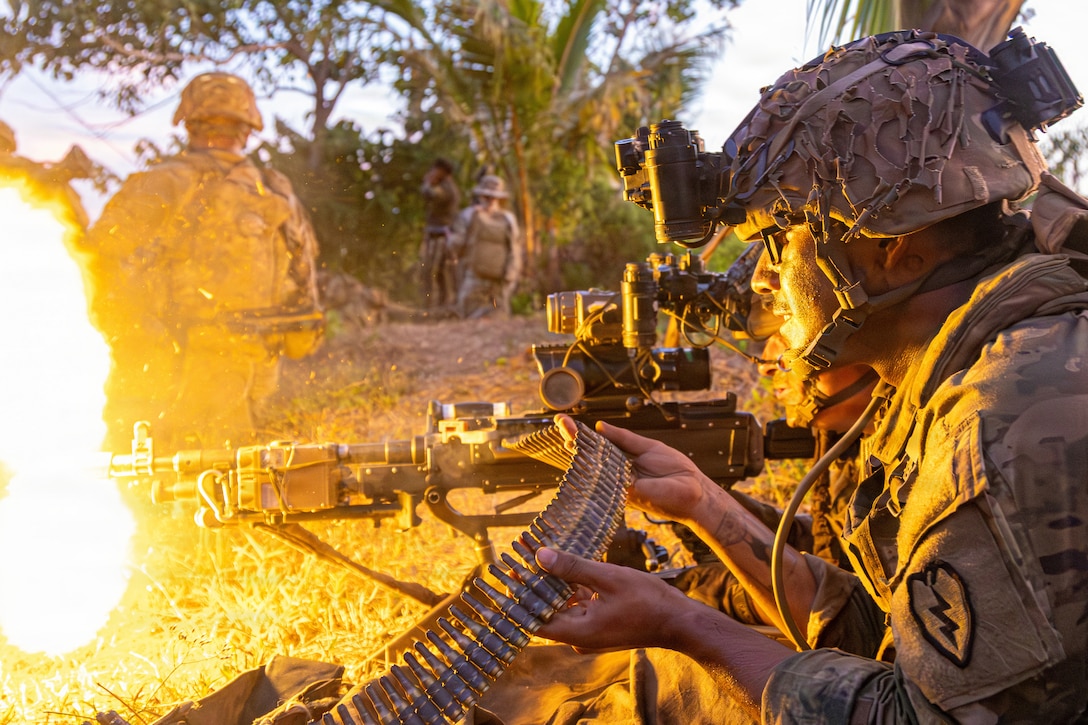 Flames shoot out the barrel of a machine gun fired by a soldier lying on his stomach, assisted by another soldier feeding a belt of cartridges into the gun. Both are wearing camouflage uniforms and helmets, and other soldiers are standing in the background near tropical plants.