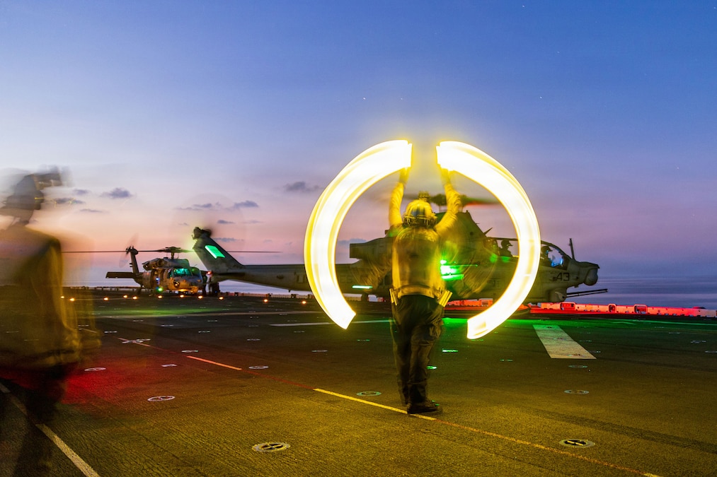 A sailor wearing a yellow shirt and vest creates circular light streaks with two handheld lights as he signals to a helicopter on the deck of a ship in low light conditions.