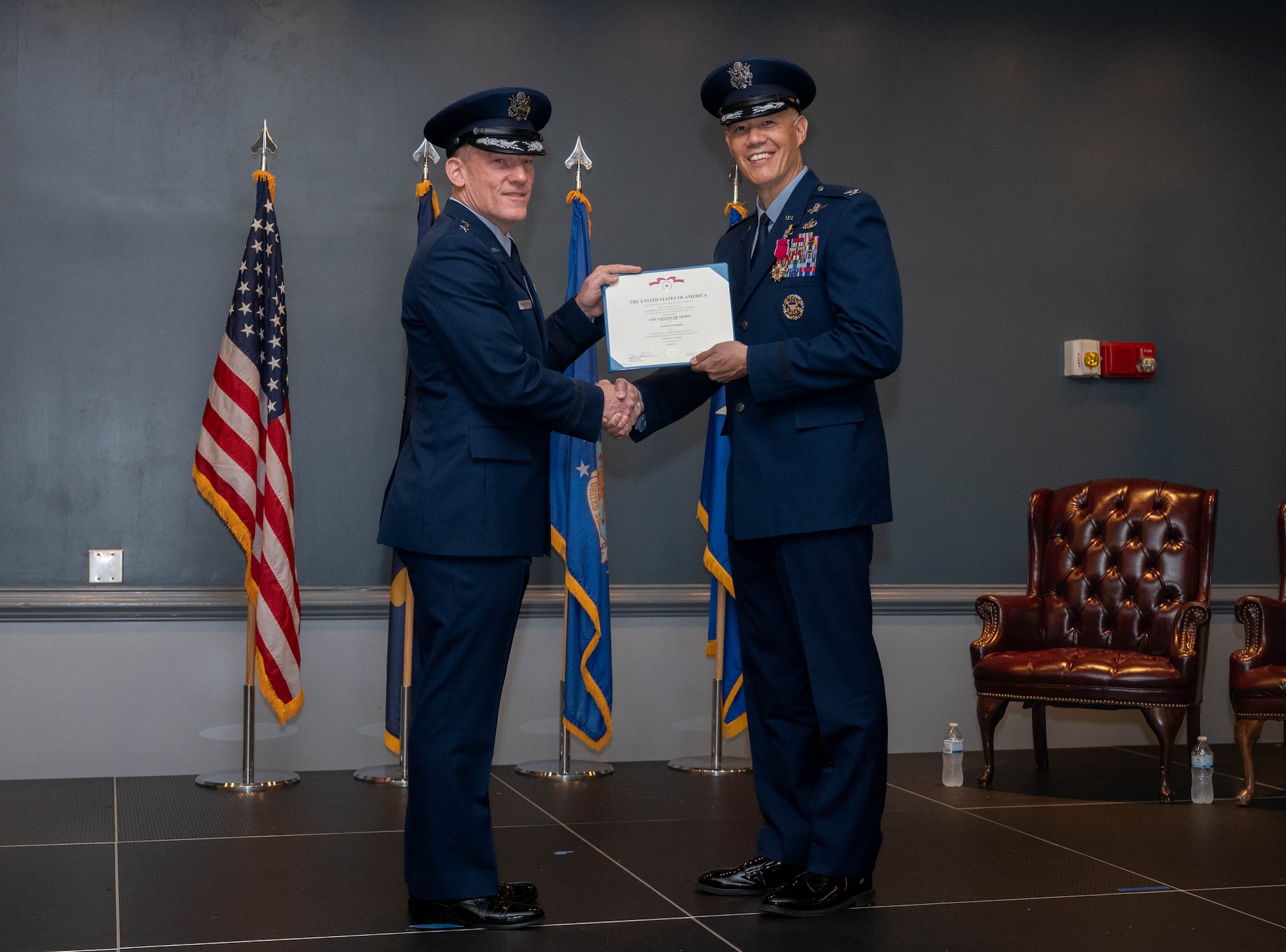 Two men in uniform hold a certificate and shake hands on a stage.