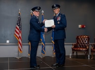 Two men in uniform hold a certificate and shake hands on a stage.