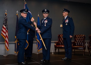 Four men in uniform stand on a stage, two holding a flag and two at attention