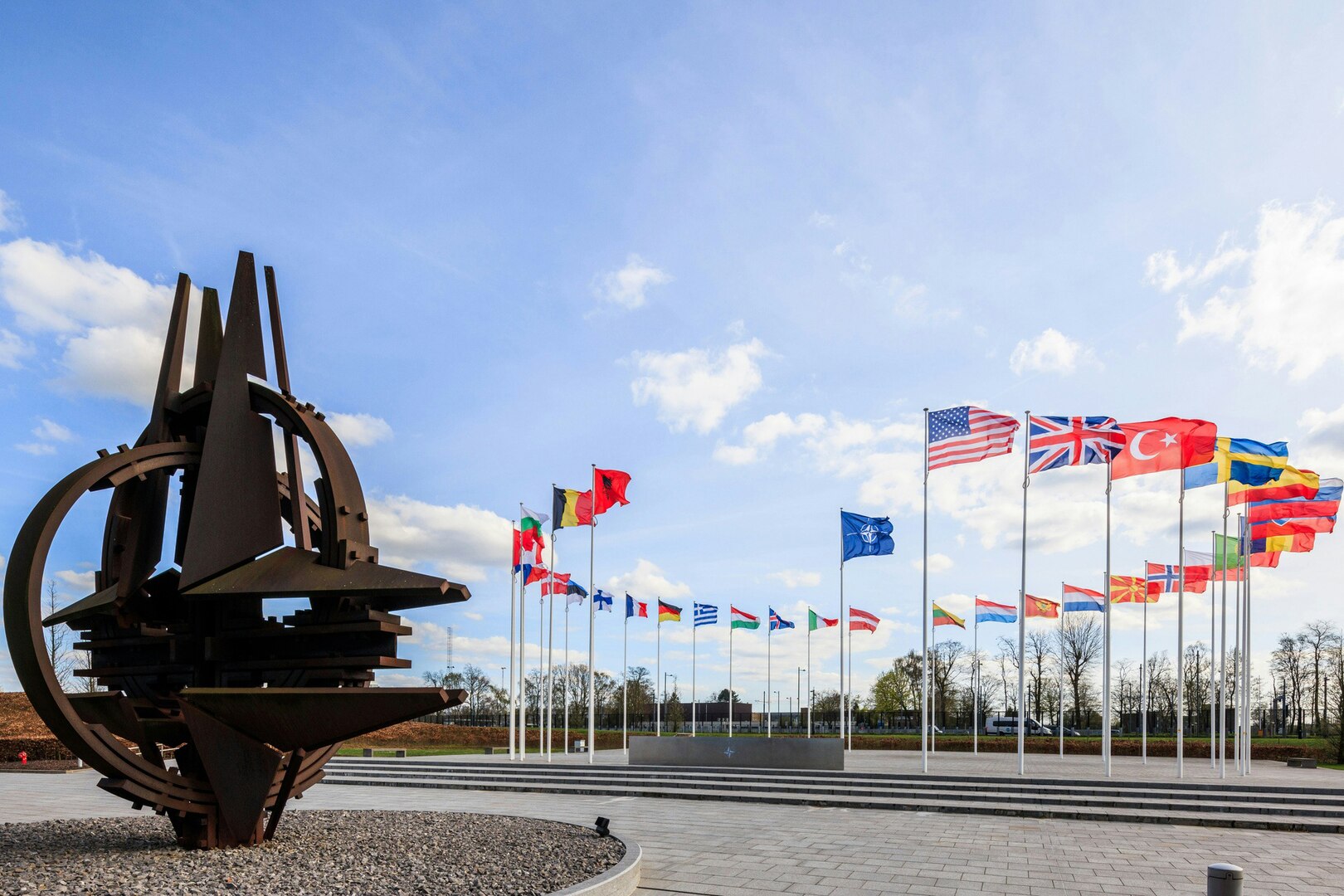 A sculpture of a stylized NATO emblem stands near a circle of national flags on a paved area.