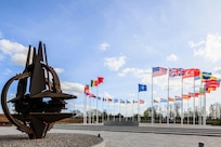 A sculpture of a stylized NATO emblem stands near a circle of national flags on a paved area.