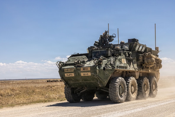 Eight-wheeled KMA Stryker vehicle rolling toward the camera in a desert setting with a service member sitting on top.
