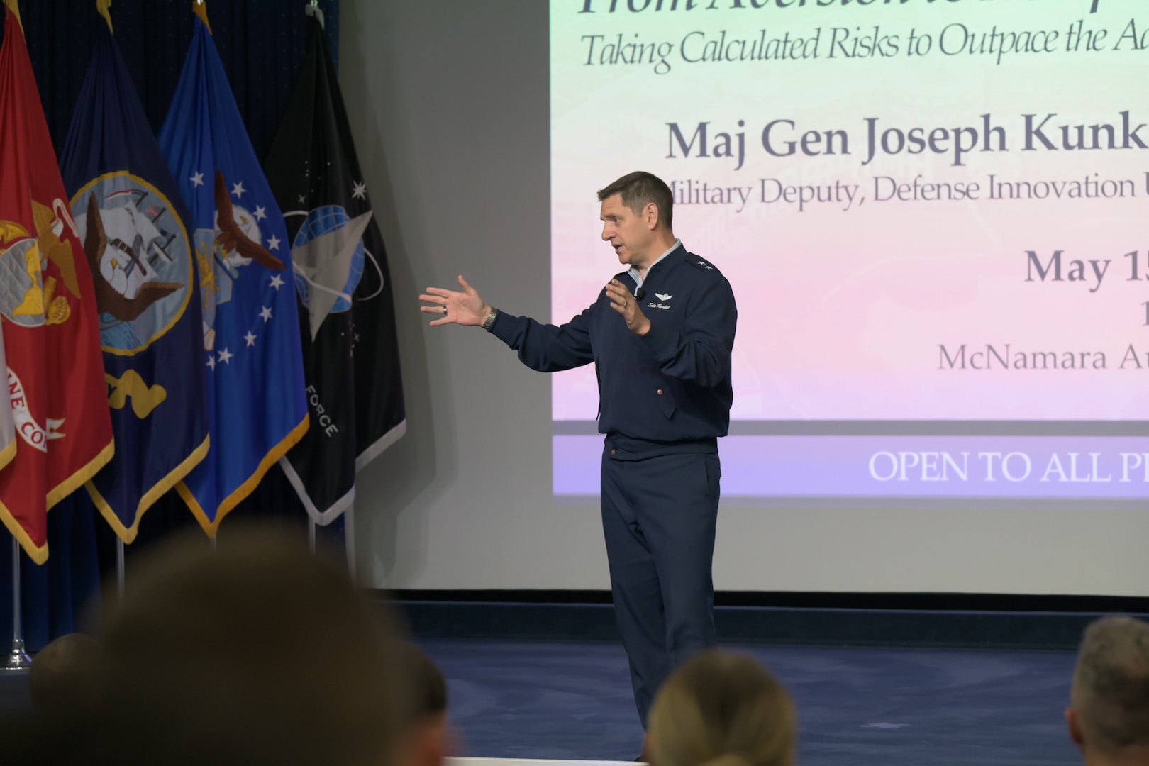 An Air Force major general stands on a stage, with military flags and a screen behind him, as he gives a talk.