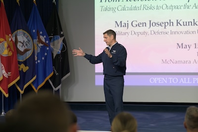 An Air Force major general stands on a stage, with military flags and a screen behind him, as he gives a talk.