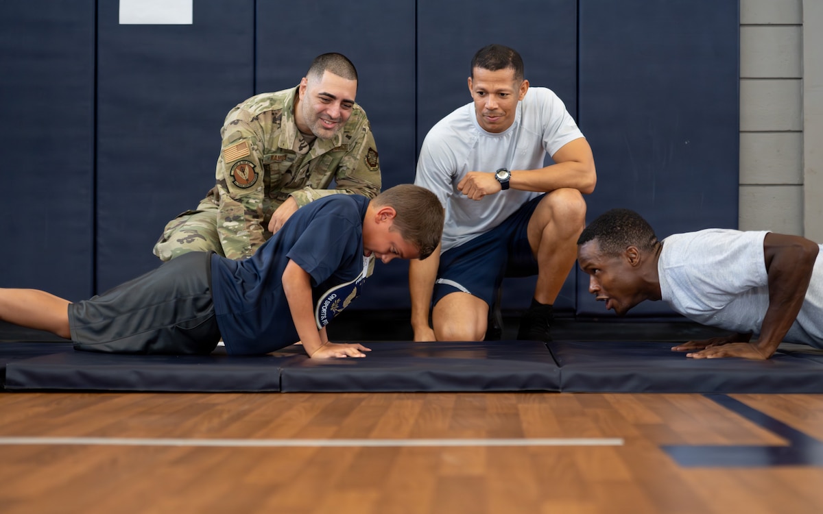 U.S. Air Force Staff Sgt. Michael Ramos, left, 6th Force Support Squadron fitness center operations manager,  Airman Domingo Galvez, center, 6th FSS fitness center specialist, and Senior Airman Marcus Higginbottom, 6th FSS fitness assessment cell specialist, motivate a military child during the push-up portion of the Presidential Fitness Test at MacDill Air Force Base, Florida, June 12, 2026.