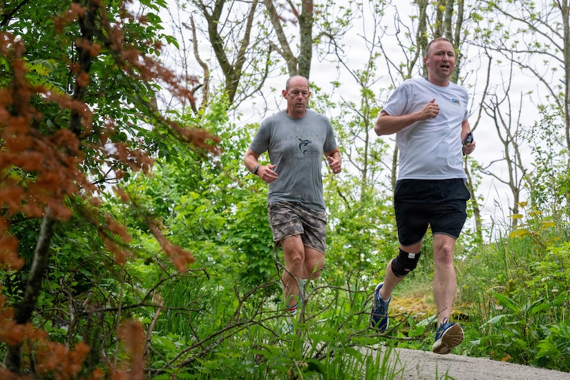 Two men in athletic attire run down a path in a forest.