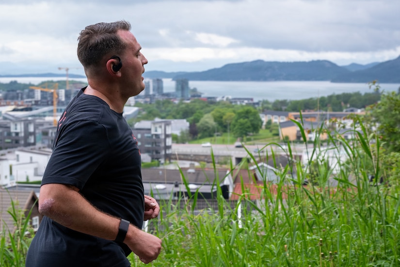 A man in athletic attire runs down a path overlooking a town on the water with mountains in the background.