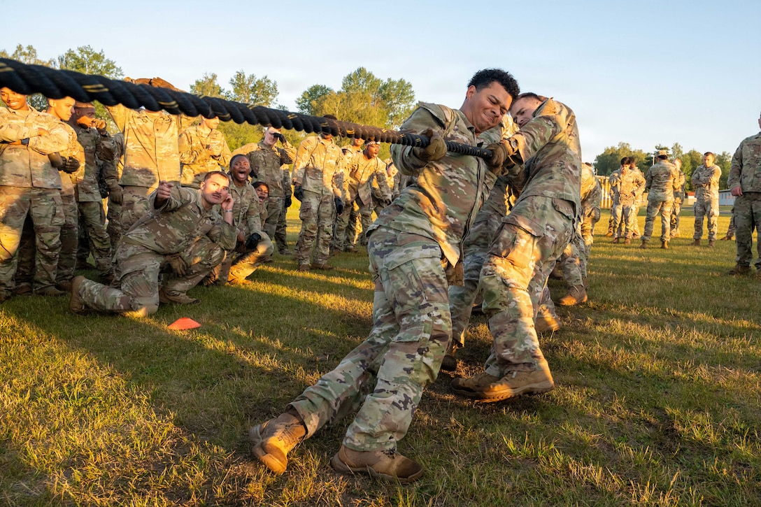Soldiers lean back while pulling hard on a thick rope, as a large group of other soldiers look on at the tug-of-war challenge.