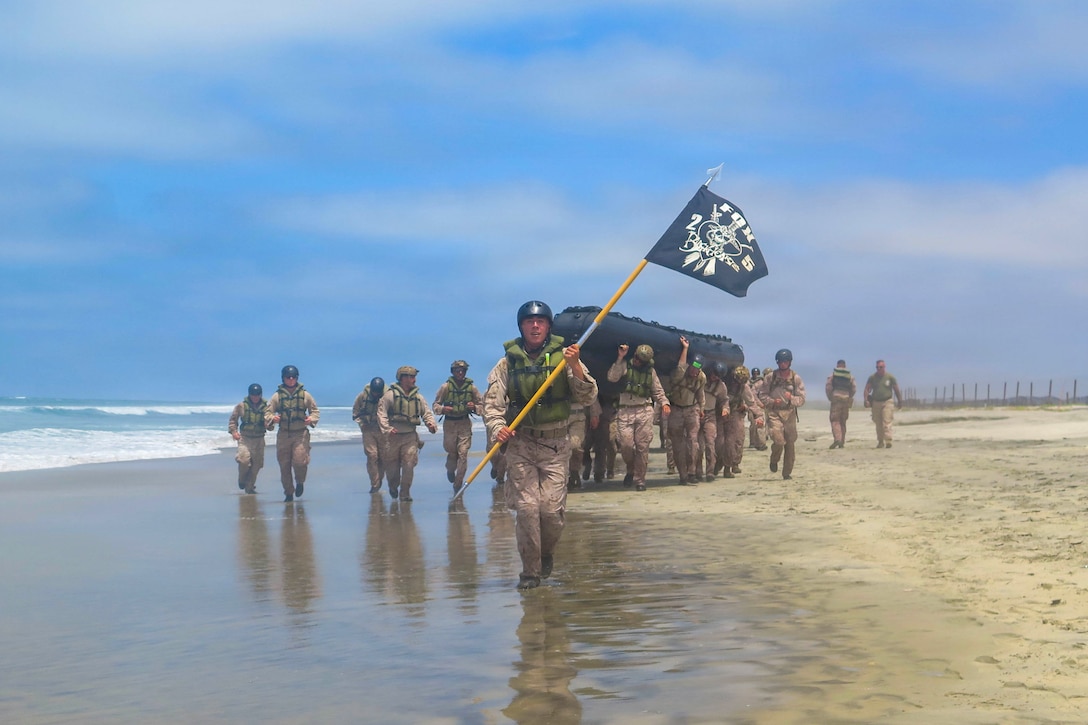 A group of Marines in camouflage and vests carries a small boat on the shore, with other Marines surrounding them. One Marine carrying a flag walks in front of them.