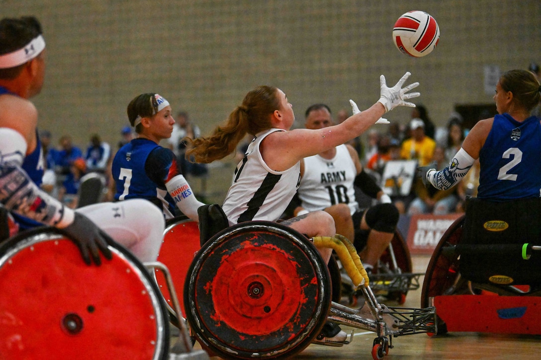 An athlete in a wheelchair reaches her arms up to grab a rugby ball that’s in the air as other players surround her.