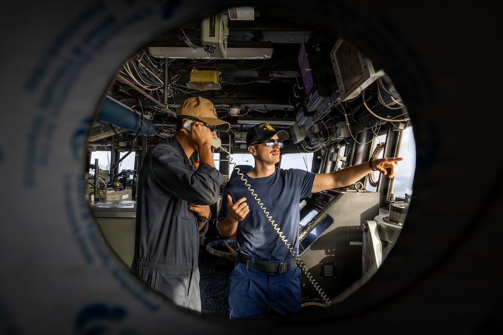 Two sailors on the other side of a small round window stand on the bridge of a ship, with one on a telephone and the other pointing out another window.