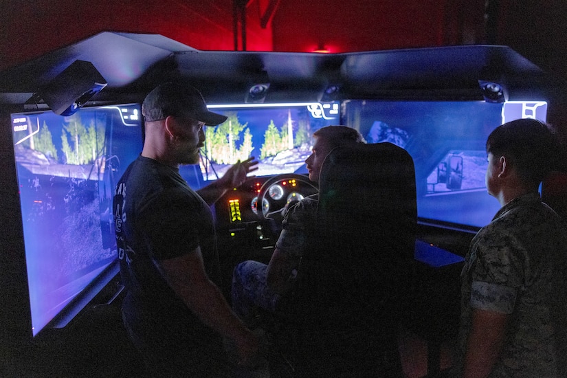 A man wearing casual attire gestures as he speaks to two other men dressed in military camouflage uniforms inside of a driving simulator.