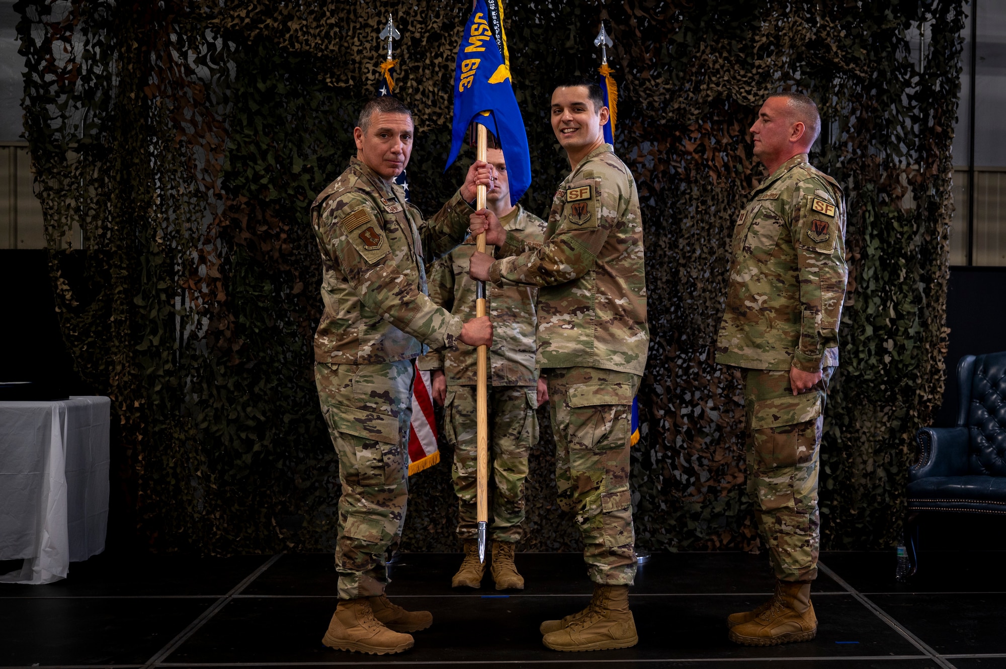 Three men in military uniforms stand on stage. Two of them hold a guidon.