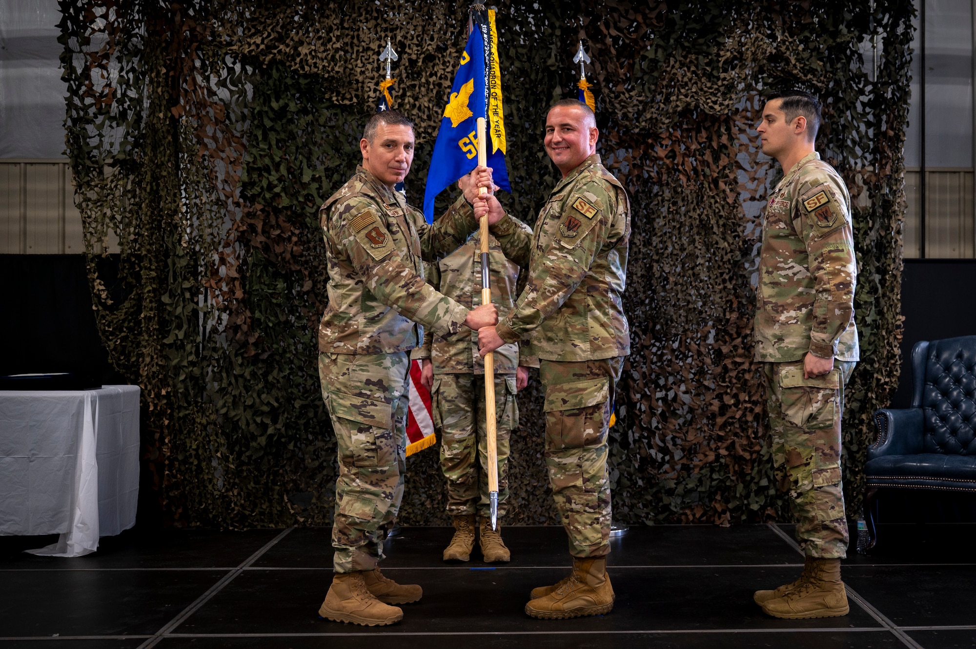 Three men in military uniforms stand on stage. Two of them hold a guidon.