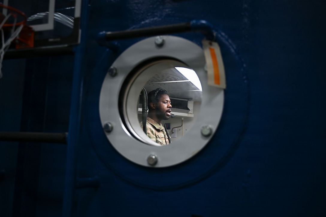 A man is seen through a circle window into the decompression chamber. He is speaking to those inside.